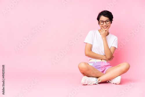 Young Vietnamese woman with short hair sitting on the floor over isolated pink background with glasses and smiling