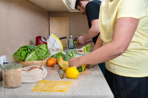 Unrecognisable woman in yellow T-shirt wrapping vegetables in beeswax cloth for storage in fridge. Zero waste and sustainable lifestyle