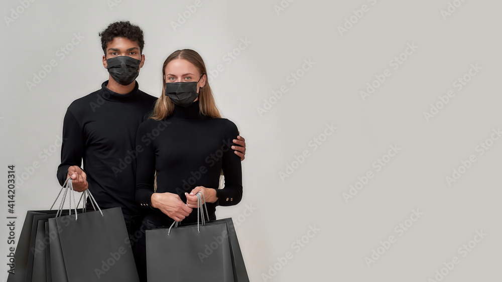Best friends wearing black outfits and protective masks looking at camera, holding shopping bags, posing together isolated over light gray background