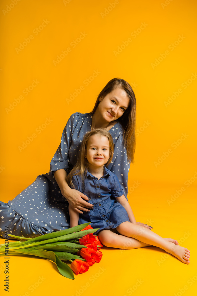 mom and daughter sit on the floor with a bouquet of red tulips and look at the camera on a yellow background with a place for text
