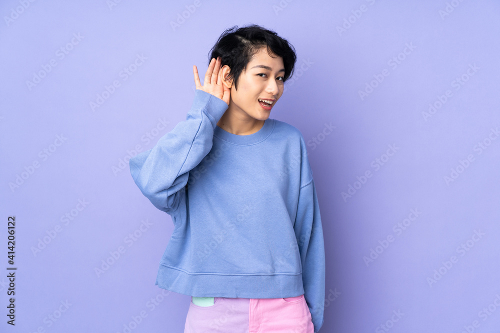 Young Vietnamese woman with short hair over isolated purple background listening to something by putting hand on the ear Young Vietnamese woman with short hair over isolated purple background listening to something by putting hand on the ear