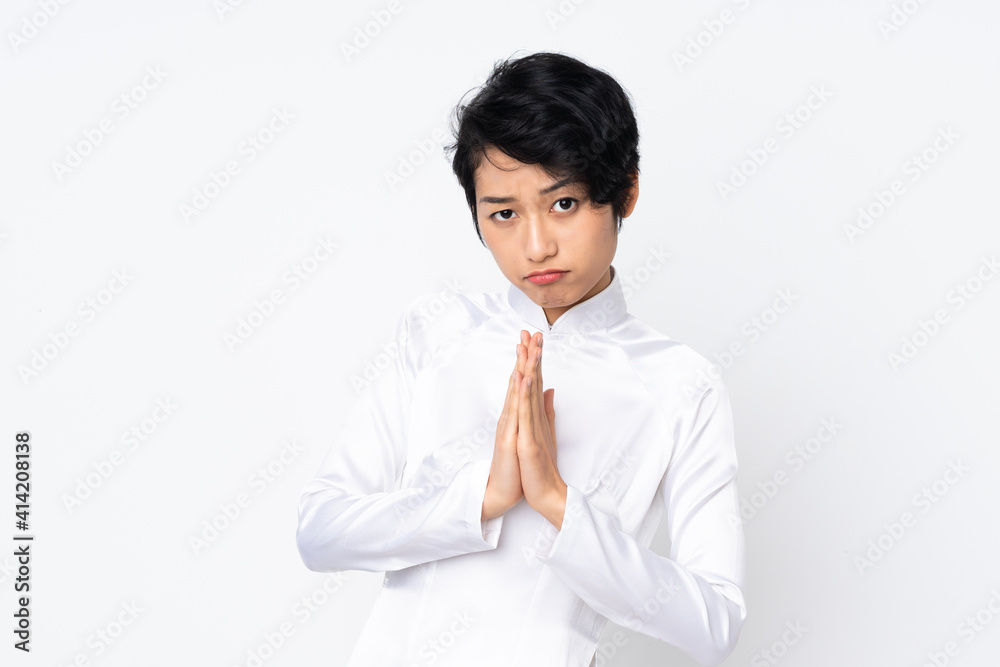 Young Vietnamese woman with short hair wearing a traditional dress over isolated white background keeps palm together. Person asks for something Young Vietnamese woman with short hair wearing a traditional dress over isolated white background keeps palm together. Person asks for something