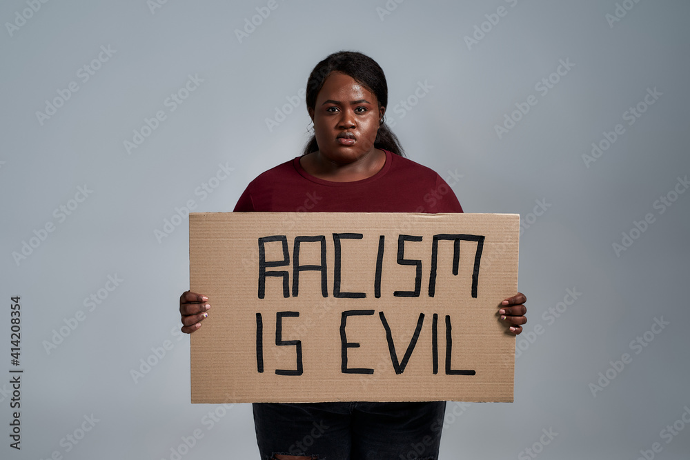 Serious plus size young african american woman in casual clothes looking at camera, holding Racism is evil banner in front of her, posing isolated over gray background Serious plus size young african american woman in casual clothes looking at camera, holding Racism is evil banner in front of her, posing isolated over gray background
