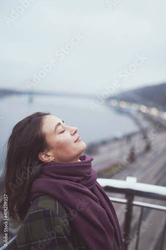 Beautiful calm young woman staying near river with closed eyes and expressing delight.