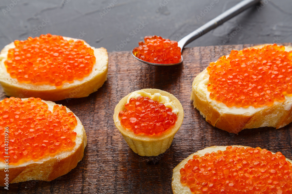 Bruschetta with butter and red caviar next to a spoon with caviar on a board on a concrete background. Bruschetta with butter and red caviar next to a spoon with caviar on a board on a concrete background.