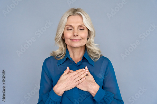 Happy mindful thankful middle aged old woman holding hands on chest meditating with eyes closed isolated on grey background feeling no stress, gratitude, mental health balance, peace of mind concept.