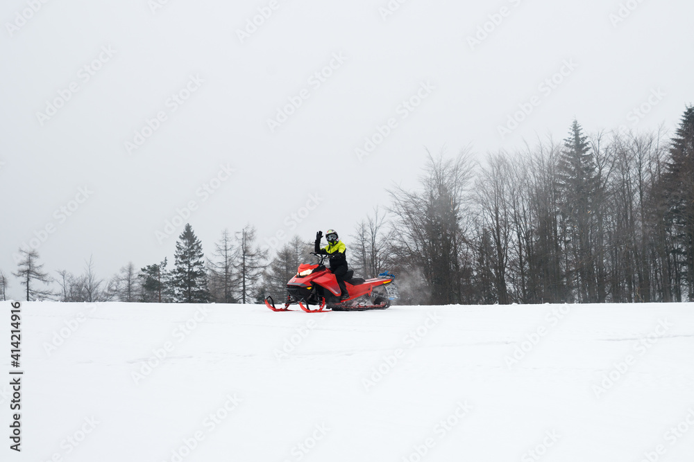 Driver is driving snowmobiles on snow field with forest background and he is waving his hand to say hello.