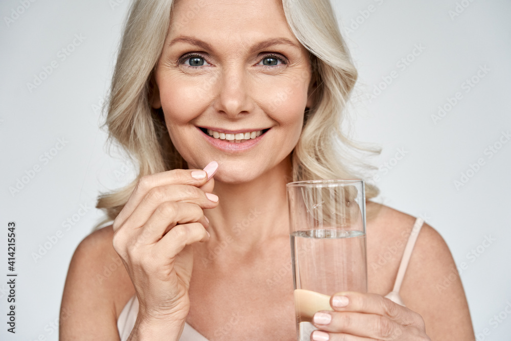 Smiling happy healthy middle aged 50s woman holding glass of water taking dietary supplement vitamin pink pill isolated on white background. Old women multivitamins antioxidants for anti age beauty. Smiling happy healthy middle aged 50s woman holding glass of water taking dietary supplement vitamin pink pill isolated on white background. Old women multivitamins antioxidants for anti age beauty.