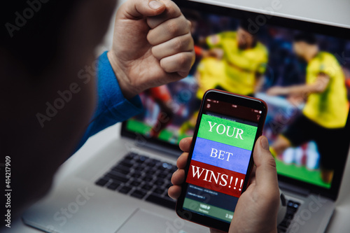 Man watching football online broadcast on his laptop and celebrate victory in betting at bookmaker's website. Focus on smartphone with message.