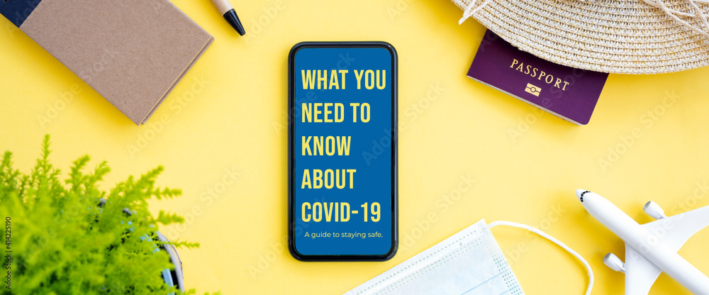 Flat lay, an aircraft model, sun hat, protective face mask, passport and a smartphone with ‘What you need to know about Covid-19, a guide to staying safe’ note, are arranged on a yellow background.