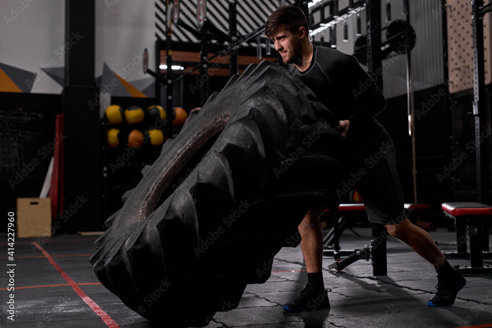 Obraz premium Side view on active man work hard - flipping tire in crossfit training, man using sportive equipment. Cross fit and workout. Strong and handsome athlete.