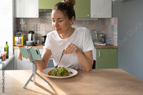 Woman eating slowly pasta with vegetarian pesto sauce, addicted to social networks, watching movie on smart phone on tripod at home kitchen. Italian cuisine, gluten free food. Slow living. 