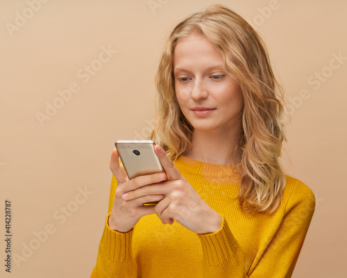 Pretty female looking social media on mobile phone standing near beige background. Natural realistic beauty, fresh face of youthful woman. Portrait of beautiful blonde in yellow jumper with cell phone