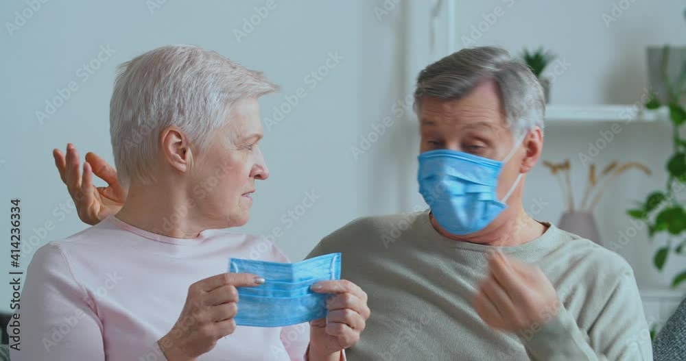 Elderly couple caucasian gray-haired man and aged woman mature family sitting together at home isolated holding put on medical protective masks discussing pandemic virus pandemic, lockdown concept