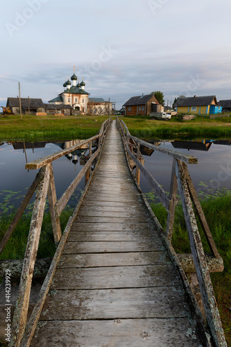 Fototapeta Naklejka Na Ścianę i Meble -  A wooden pathway across a pond connects two parts of Vershinino village, the center of Kenozersky National Park. Church of the Assumption of the Blessed Virgin Mary is seen on the far shore of the pon