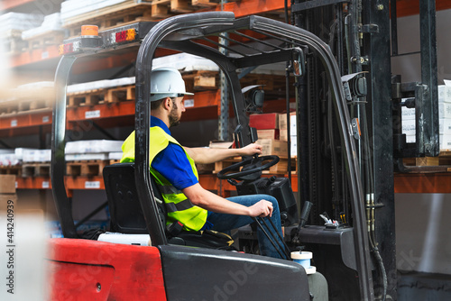 Caucasian male in a uniform and helmet driving the forklift through a storage room in a factory