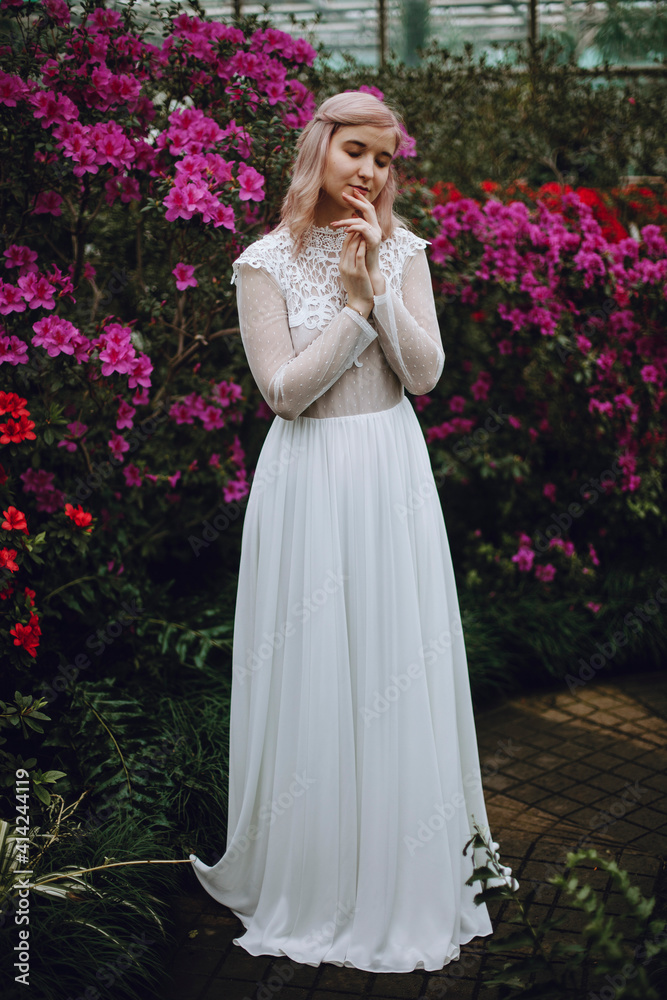 young girl in a white vintage dress stands with closed eyes in a flower greenhouse