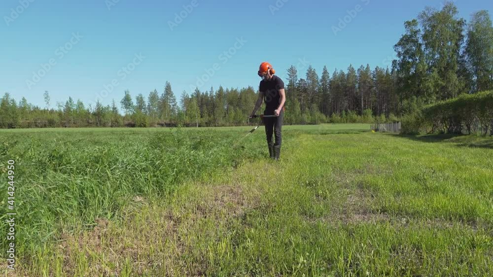 Cute young Caucasian boy mowing native grass with petrol lawn trimmer in field. Gardening care tools and equipment. Process of lawn trimming with hand mower, face, eyes and ears protection Cute young Caucasian boy mowing native grass with petrol lawn trimmer in field. Gardening care tools and equipment. Process of lawn trimming with hand mower, face, eyes and ears protection
