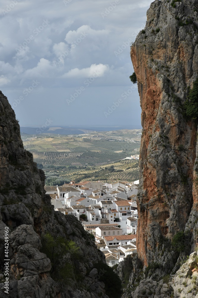 Partial view of the white village of Zahara de la Sierra, Spain, clipped by limestone cliffs from the nearby Garganta Verde canyon, endless hills of olive groves and a stormy sky in the distance