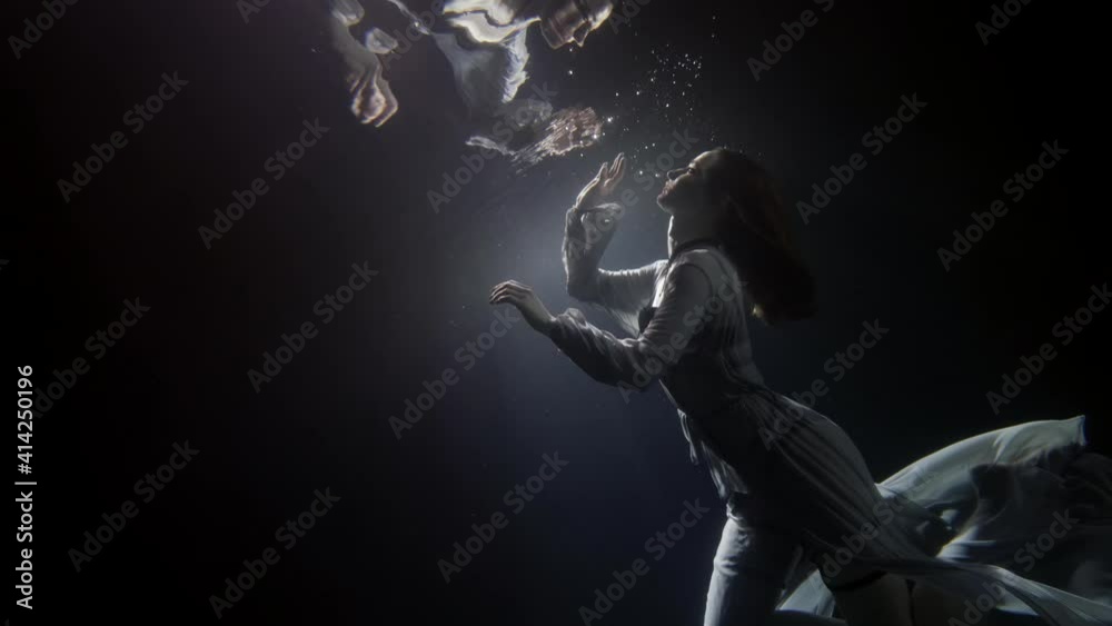 A beautiful woman in a long flowing dress floats under the water on a dark background, she swims to the surface of the water to her reflection, creating a lot of bubbles.