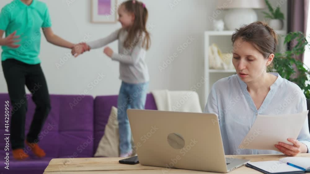 woman studies with notes at the computer, at home. children jump and have fun on the couch and want to attract the attention of the mother. the girl is focused on her studies and ignores the children woman studies with notes at the computer, at home. children jump and have fun on the couch and want to attract the attention of the mother. the girl is focused on her studies and ignores the children