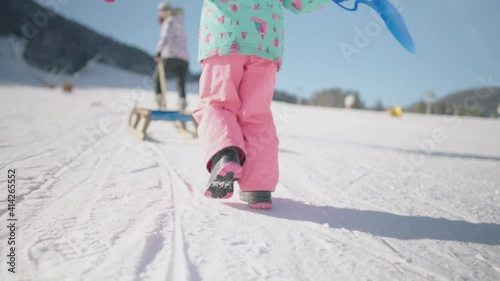 Slow motion close up - Little girl walking uphill in snow park to go sledding. Fun family time