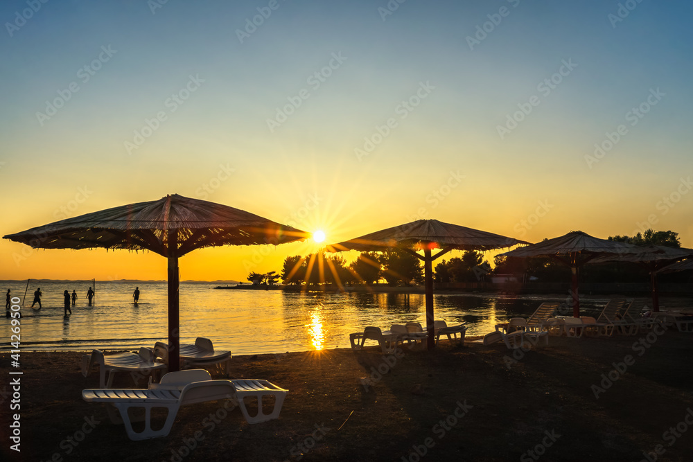 Silhouette of people playing volleyball at sunset on the Adriatic Sea beach with defined Sunstar between straw umbrellas, holidays in Zaton, Croatia