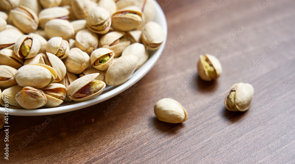 Closeup of pistachio nuts on white porcelain plate on wooden table, selective focus