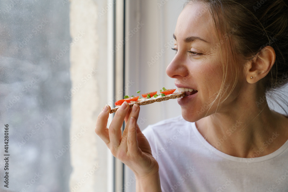 Happy woman eating rye crisp bread with creamy vegetarian cheese tofu, cherry tomato and rucola micro greens, sitting at home and looking at window. Healthy food, gluten free, diet concept.  - obrazy, fototapety, plakaty 