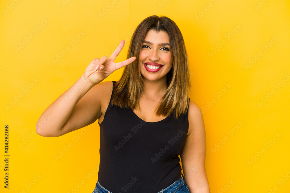 Young indian woman isolated on yellow background showing victory sign and smiling broadly. Young indian woman isolated on yellow background showing victory sign and smiling broadly.