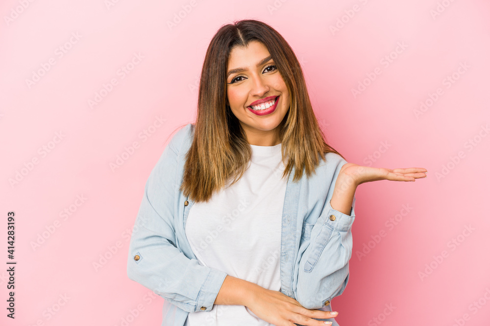 Young indian woman isolated on pink background showing a copy space on a palm and holding another hand on waist.