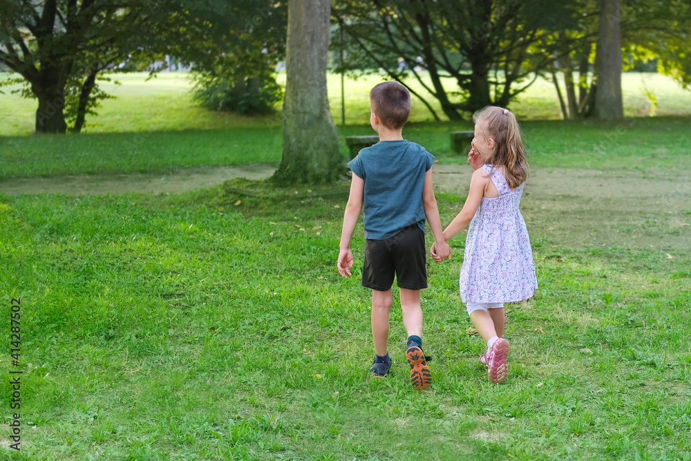 children walk in a summer park, a boy in shorts and a girl of 6-7 years old in dress are walking together on playground, holding hands, concept of active lifestyle, children's friendship