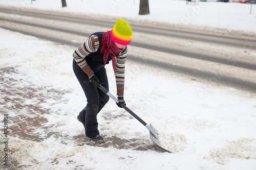 Snow removal, woman in a colorful cap with a snow shovel, clearing snow from the pavement in front of the house, winter attack