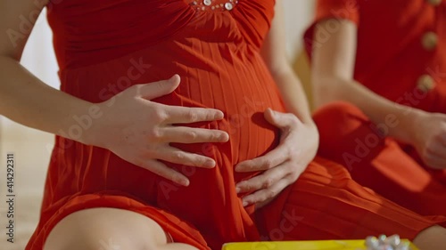 Close-up of slim pregnant Caucasian woman and unrecognizable teenage girl caressing belly in red dress. Happy carefree mother and daughter at home indoors. Pregnancy and family concept.