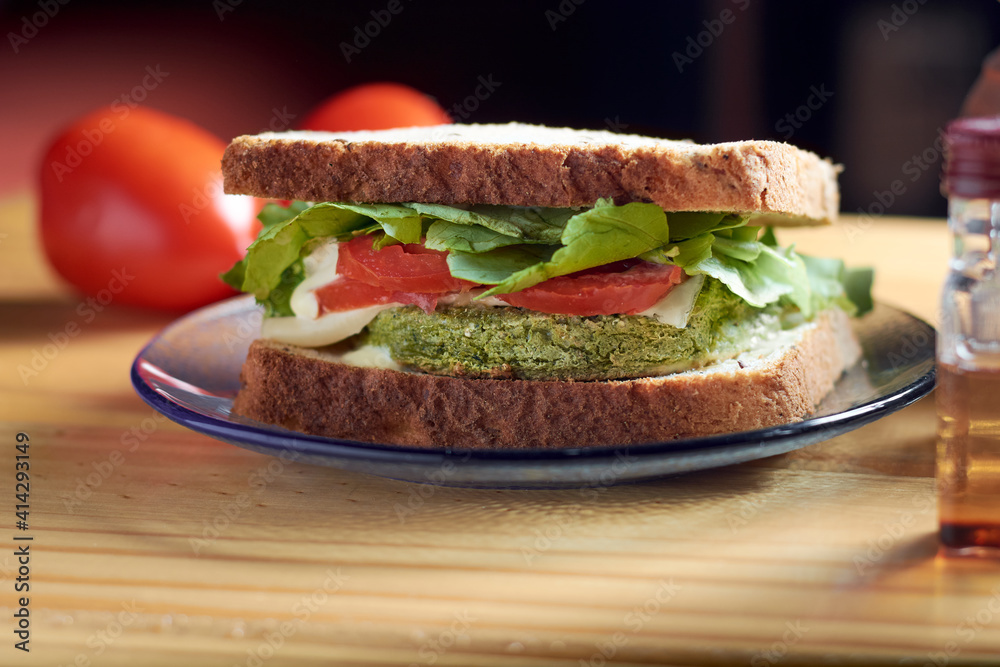 A closeup of vegan sandwich with medallion of spinach and carrot, tomato, lettuce, vegan cheese and mayonnaise on rustic wooden table. Bread with seeds. A closeup of vegan sandwich with medallion of spinach and carrot, tomato, lettuce, vegan cheese and mayonnaise on rustic wooden table. Bread with seeds.
