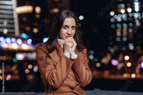Close up portrait of a lovely pleasant dark-haired lady in a coat posing against the backdrop of the bright lights of the night city looking at the camera from the belts. Female beauty concept