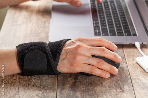 Chronic trauma to the wrist joint  in people using computer mouse may lead to disorders that cause inflammation and pain. A woman working on desk uses wrist support brace and ergonomic vertical mouse photo