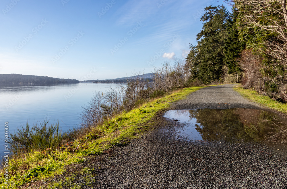 View of calm water, blue sky, and mountains from the Galloping Goose Trail in East Sooke, Vancouver Island View of calm water, blue sky, and mountains from the Galloping Goose Trail in East Sooke, Vancouver Island