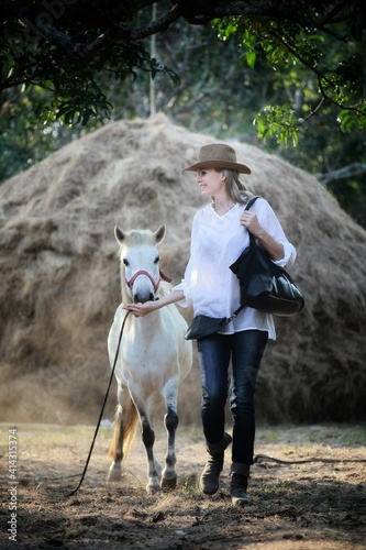 A girl in a white shirt wearing a cowboy hat with a white horse on the background of a haystack at sunset