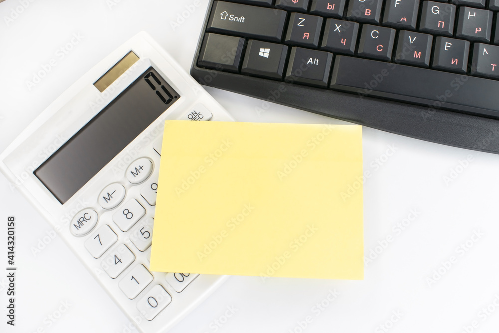 On a white background a keyboard and a white calculator with a yellow blank sheet for notes On a white background a keyboard and a white calculator with a yellow blank sheet for notes