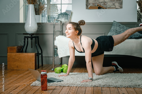 Young woman goes in for sports at home, training online. The athlete swings leg , watches a movie and studies from a laptop on background bed, vase, carpet