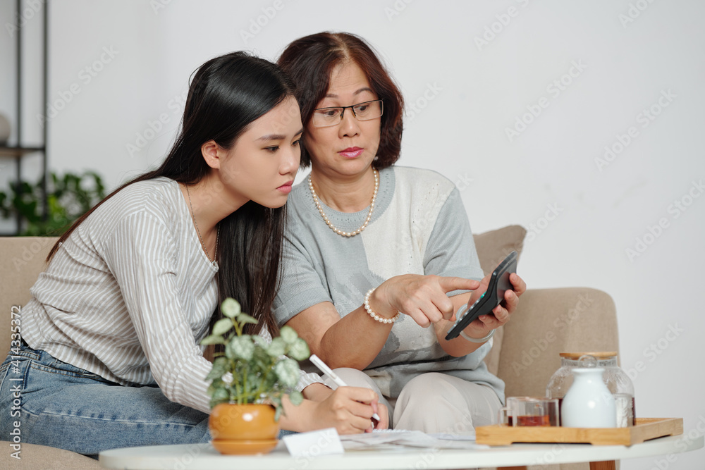 Shocked senior woman showing calculator with risiculously big number to her daughter after they summed up all expenses