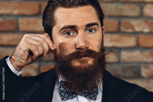 Photography Portrait of a handsome lovely man wearing a suit, hussar mustache and beard and posing against a brick wall and touching his mustache