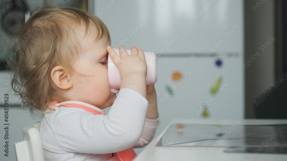 Side view little cute white girl blonde with pink bib drinks kefir from pale pink glass and looks at camera. Mouth in kefir or yogurt. Baby wear white shirt or bodysuit. Side view little cute white girl blonde with pink bib drinks kefir from pale pink glass and looks at camera. Mouth in kefir or yogurt. Baby wear white shirt or bodysuit.