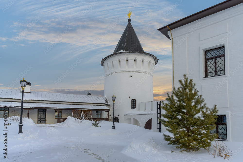Winter in the Tobolsk Kremlin (Russia). One of the round white towers in the evening against the backdrop of a sunset yellow-orange blue sky. young pine tree, lanterns and old buildings of the Kremlin