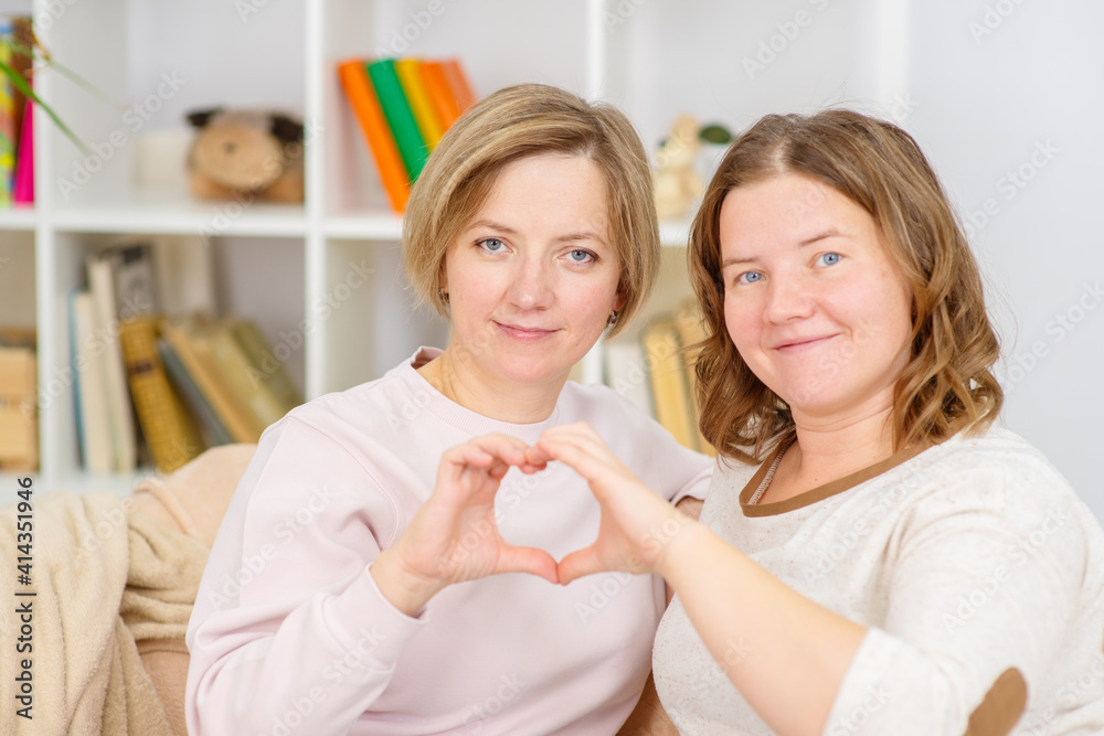 Two young women hug at home on the couch and make a heart out of their hands. Gay lesbian couple at home. Lesbian couple concept