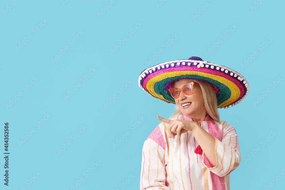Mature Mexican woman in sombrero hat pointing at something on color background