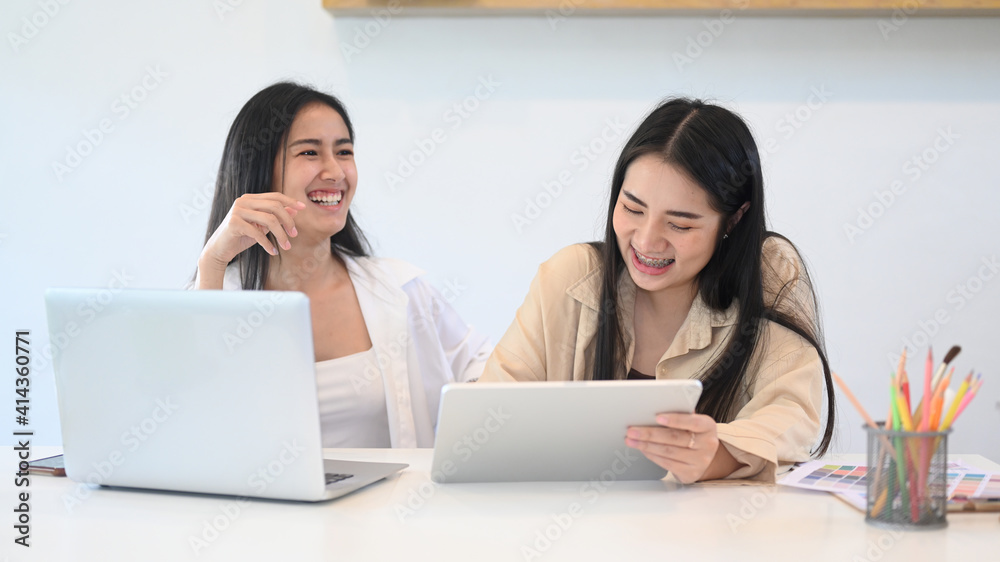 Two young woman designer in casual wear laughing while working with laptop and digital tablet at modern office. Two young woman designer in casual wear laughing while working with laptop and digital tablet at modern office.
