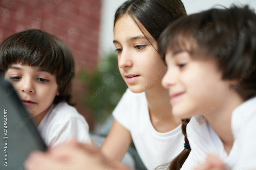 Real friendship. Close up of pretty teenage latin girl using digital tablet. Sister spending time with her two little brothers, lying on the bed at home
