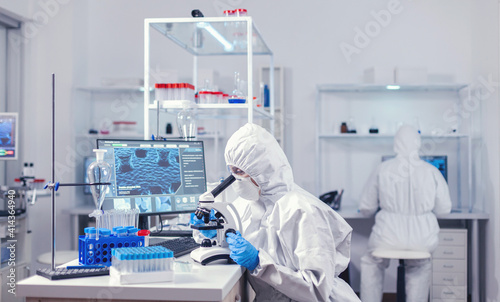 Woman medical scientist sitting at workplace in laboratory looking through microscope. Chemist researcher during global pandemic with covid-19 checking sample in biochemistry lab